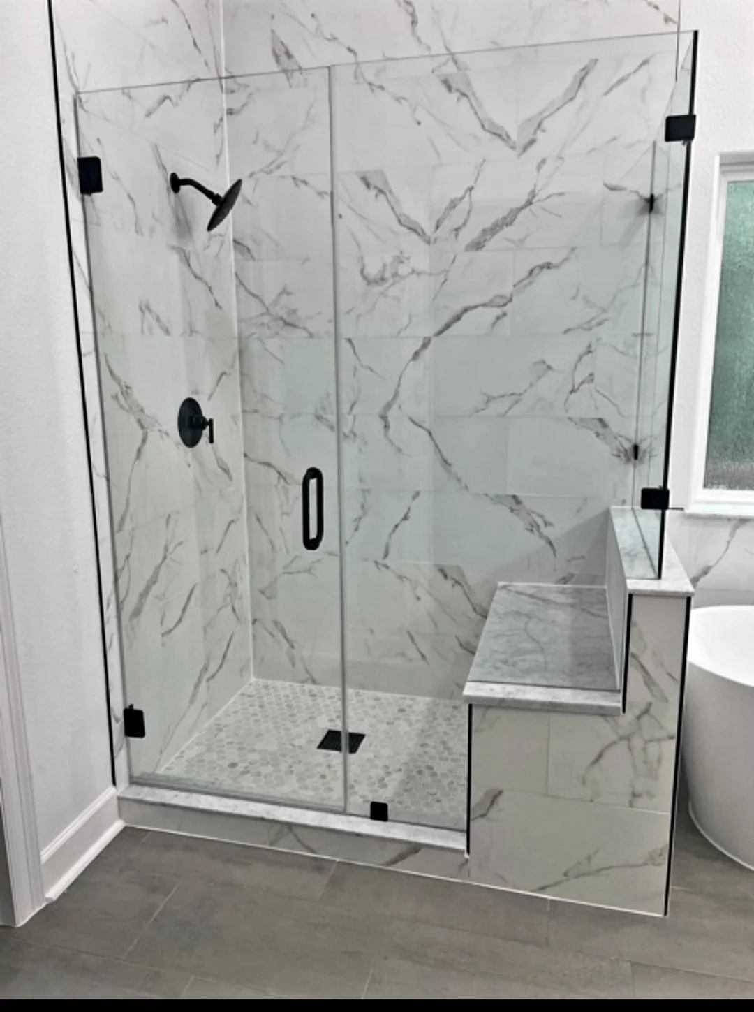 Modern walk-in shower with glass walls and black fixtures, marble tile walls, and a built-in marble bench.