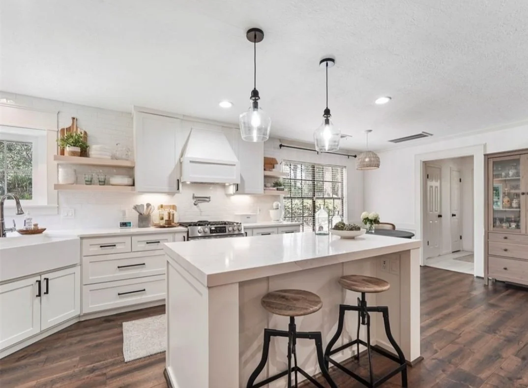 Modern white kitchen with island, wooden bar stools, pendant lights, and large window showing outdoor greenery.