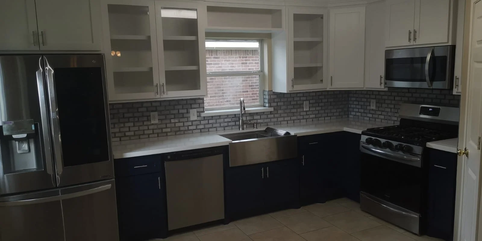 Modern kitchen with white upper cabinets, black lower cabinets, stainless steel appliances including a refrigerator, microwave, and oven, a brick backsplash, a window above the sink, and beige tiled floors.