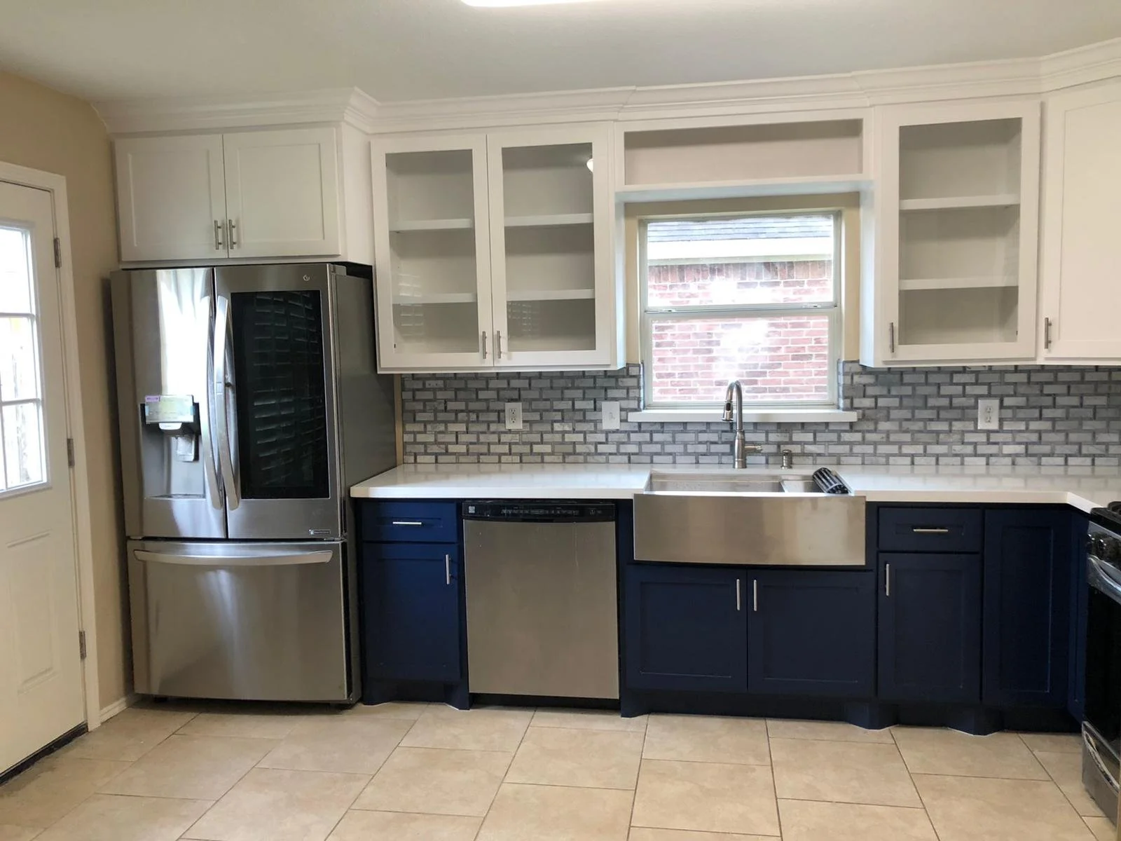 Kitchen with white upper cabinets, dark blue lower cabinets, a stainless steel refrigerator, a farmhouse sink, and a gray brick backsplash.