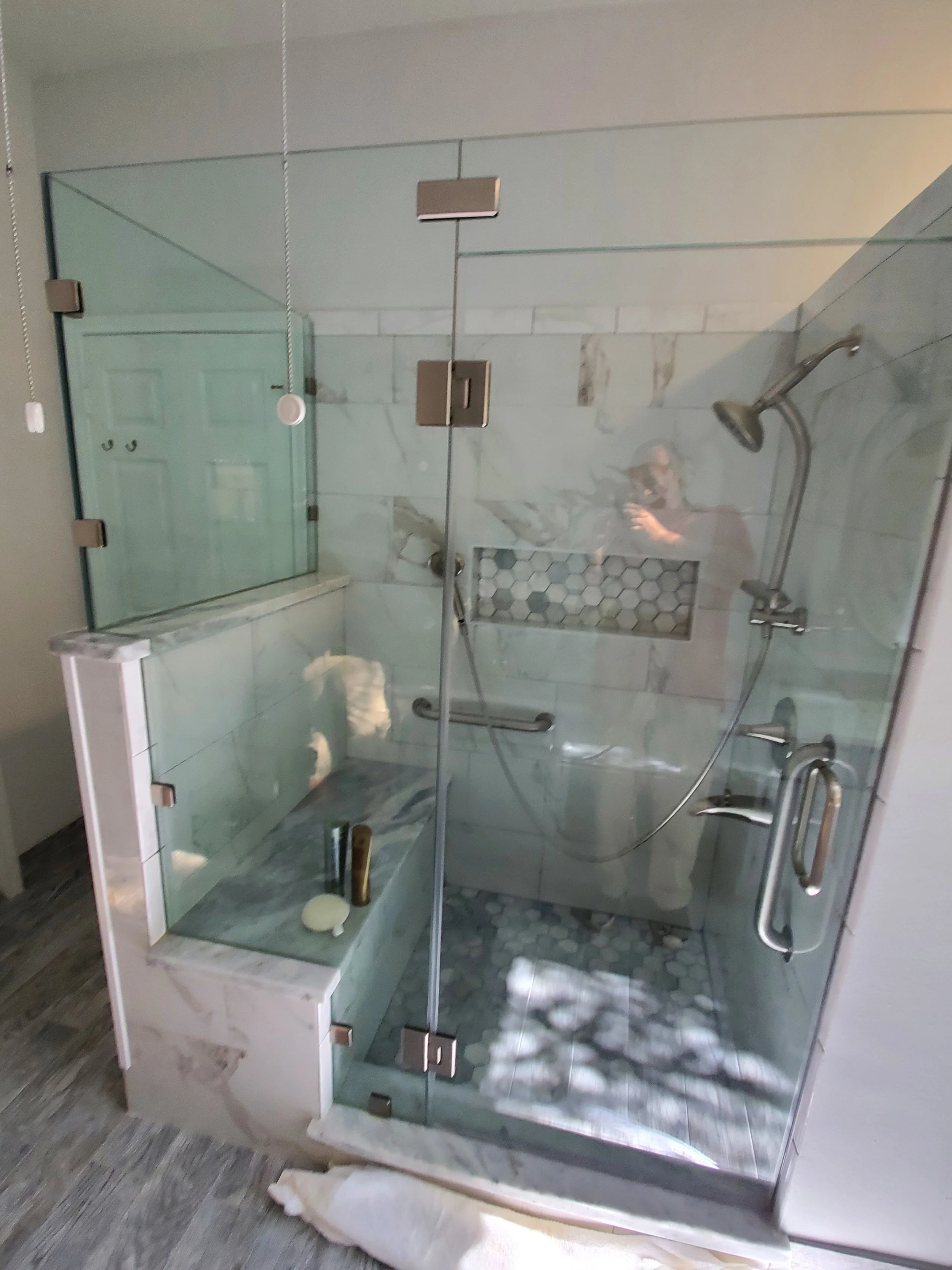 A glass shower enclosure with metal hinges and handles, showing a shower head, faucet, and tiled interior with a small built-in shelf holding bathroom supplies. The photo captures a reflection of a person taking the picture.