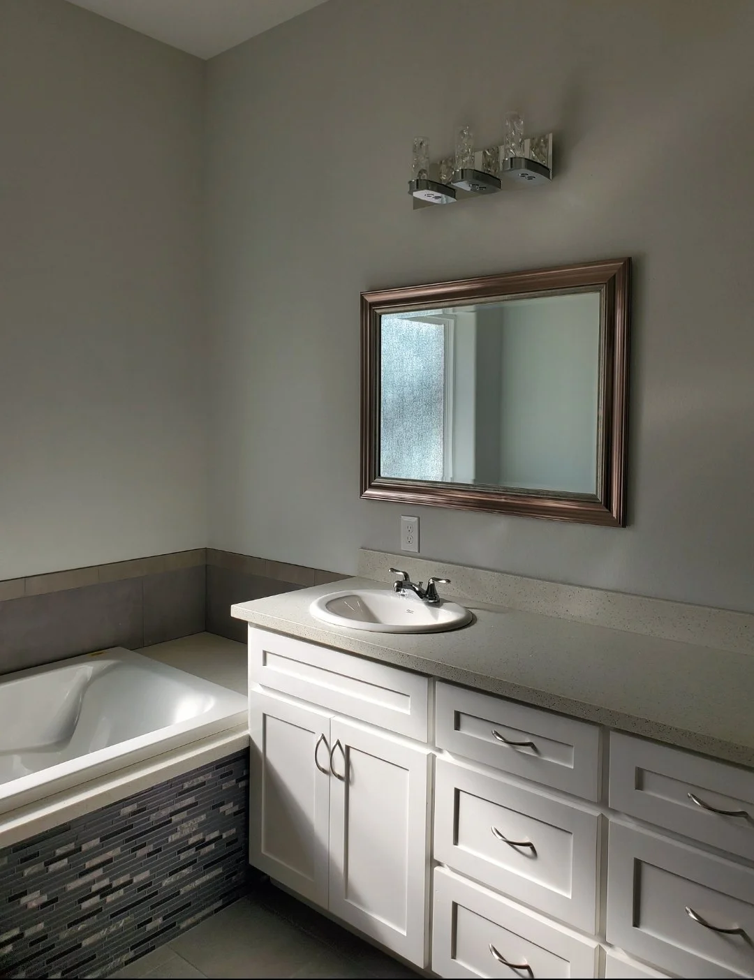 A bathroom with a white vanity, a sink, a mirror, and a bathtub with a tile surround. The bathroom has a light-colored countertop and a modern light fixture above the mirror.