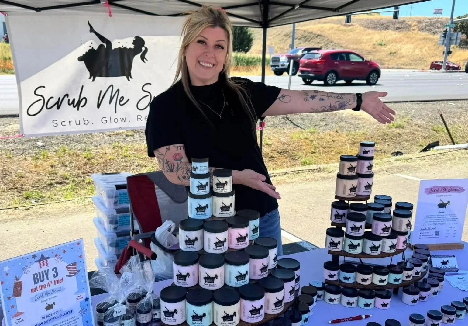 A woman smiling under a black canopy at a booth selling scented scrub jars with a banner reading 'Scrub Me Sweet' and 'Scrub. Glow. Repeat.' in an outdoor setting with cars on a highway behind her.