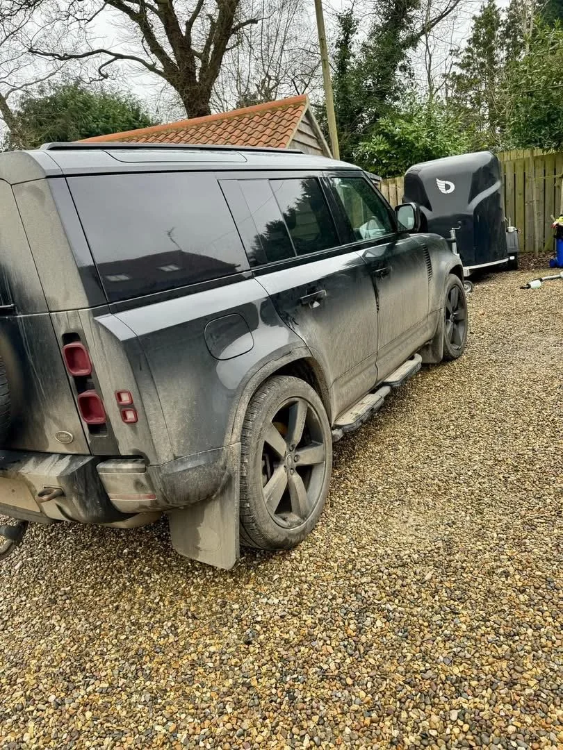 A black Land Rover SUV with dirty, dusty exterior, parked on a gravel driveway next to a black trailer and surrounded by trees and a wooden fence.