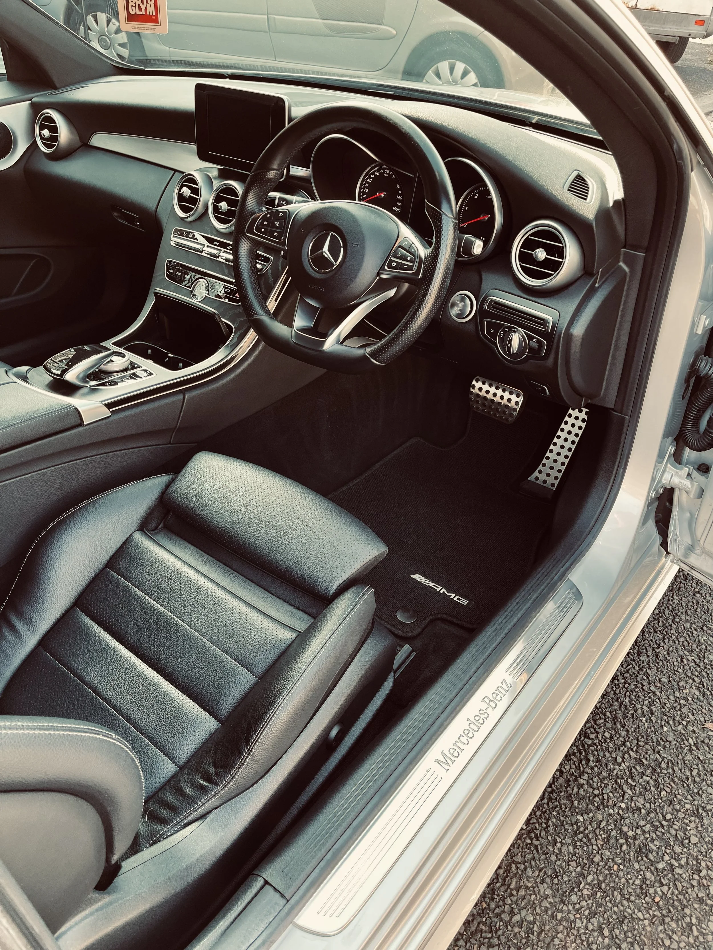 The interior of a Mercedes-Benz car showing the steering wheel, dashboard, center console, and front seat, with a view of the street outside through the windshield.