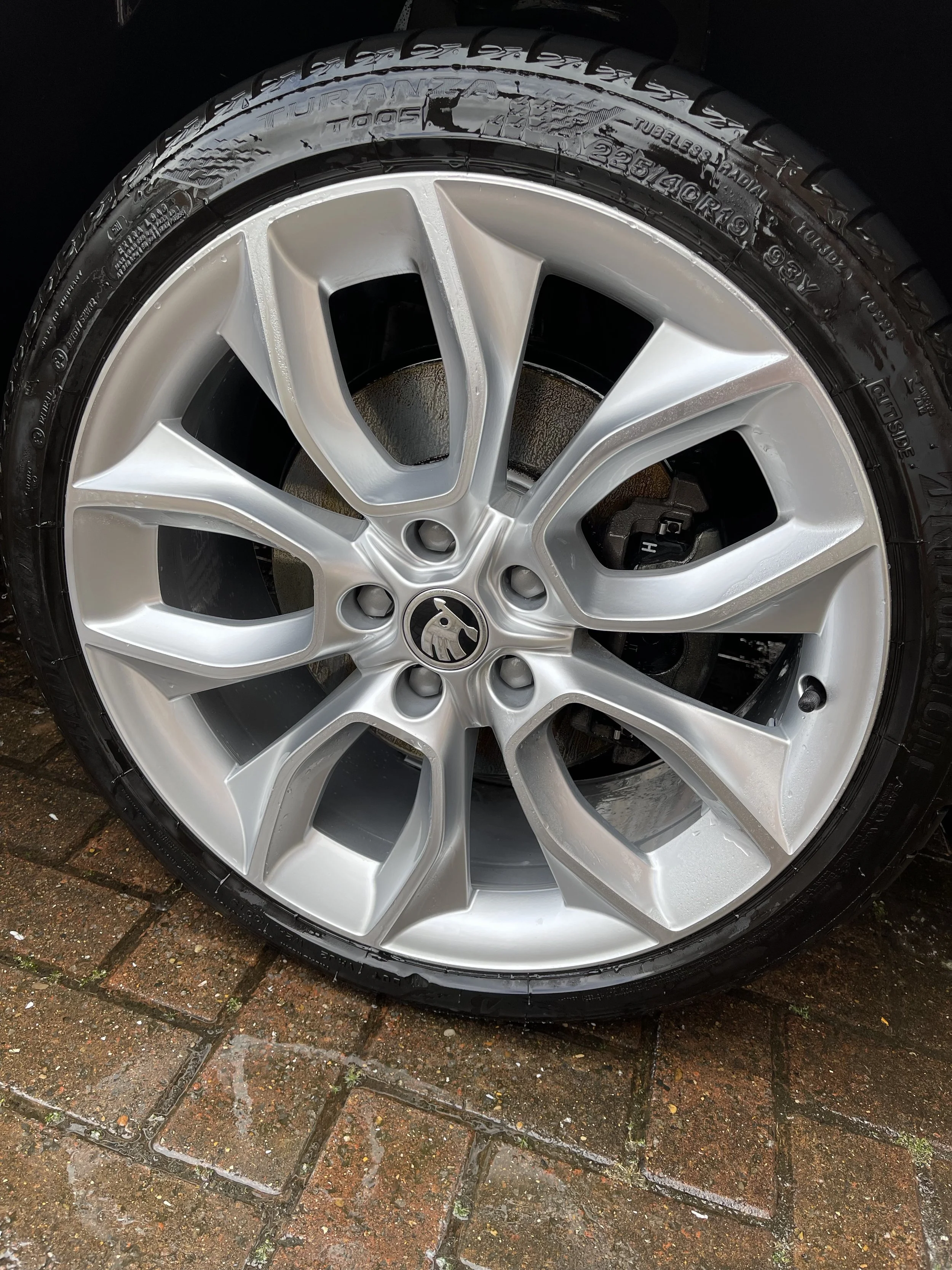 Close-up of a silver alloy car wheel with a tire mounted on it, resting on a wet brick pavement.