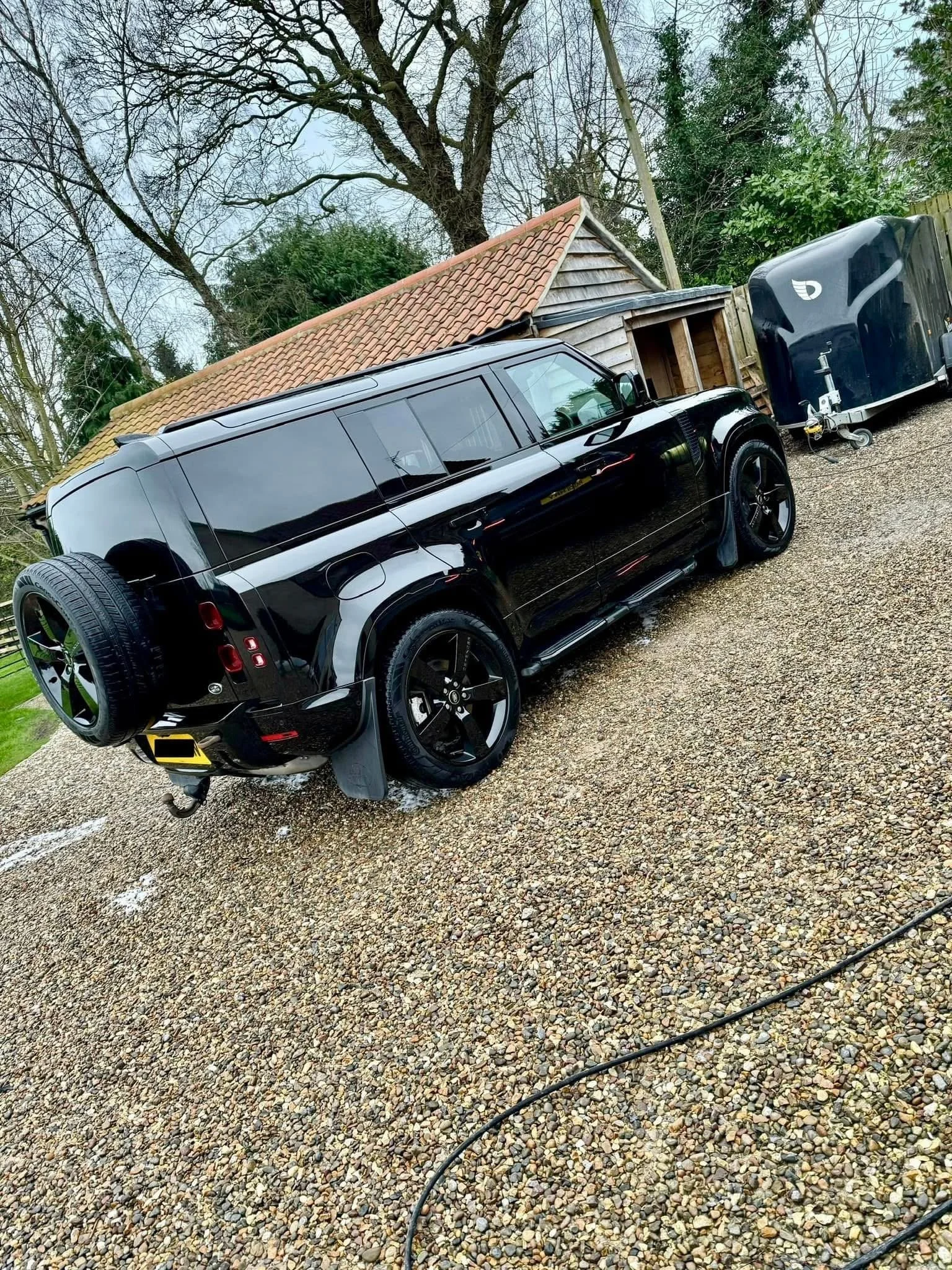 A black luxury SUV parked on a gravel driveway, attached to an enclosed trailer, beside a wooden shed with a tiled roof, surrounded by trees and greenery.