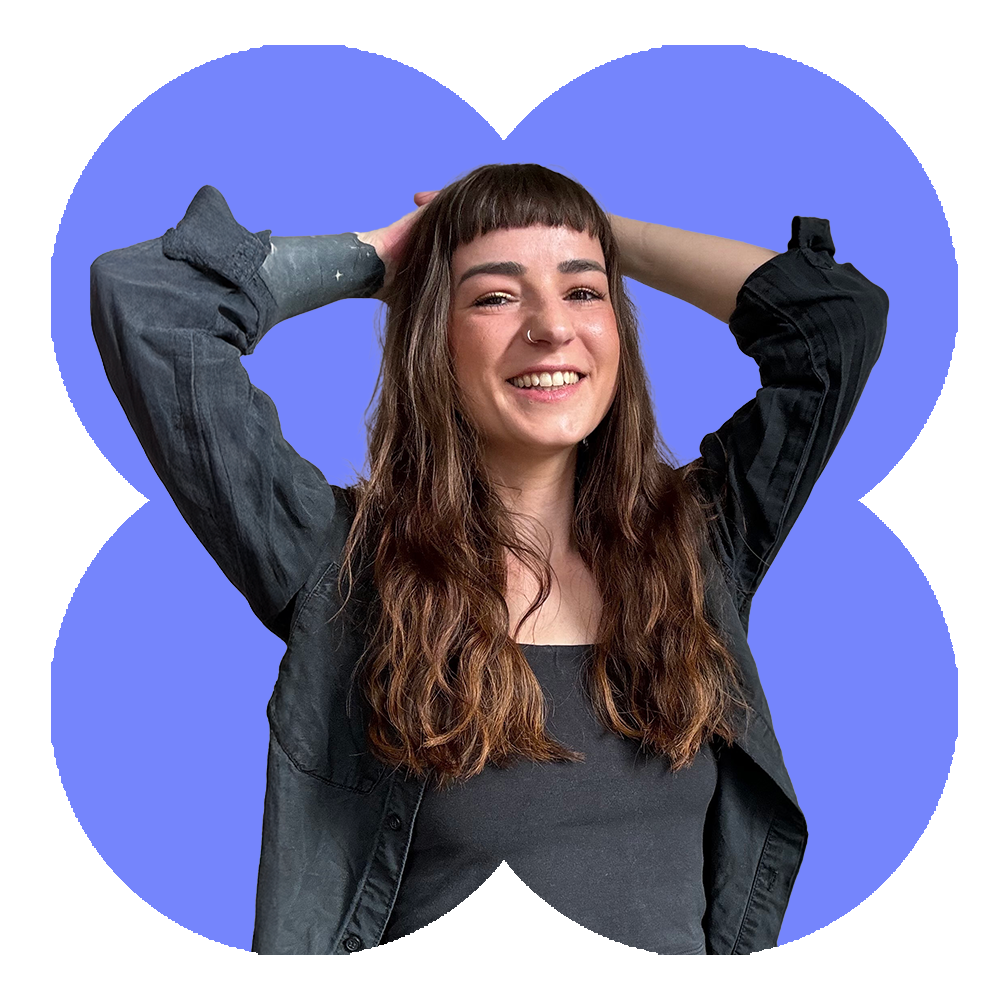 A woman with long wavy brown hair, smiling with her arms raised behind her head.