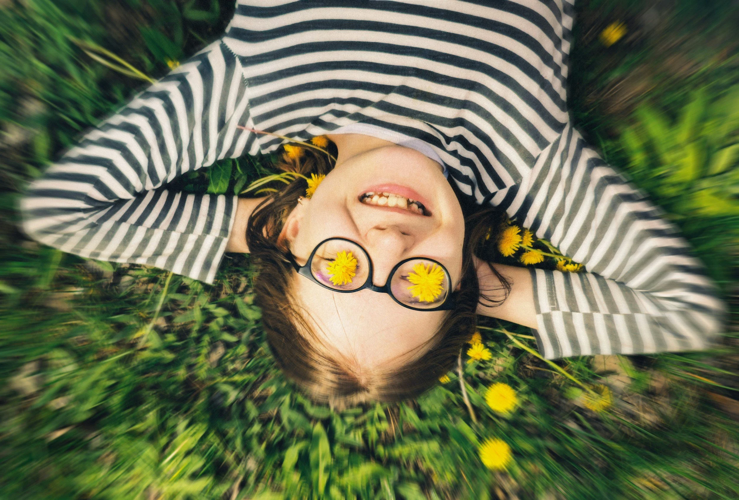 Woman lying on grass among yellow flowers, smiling with glasses reflecting yellow flowers, wearing a striped shirt.