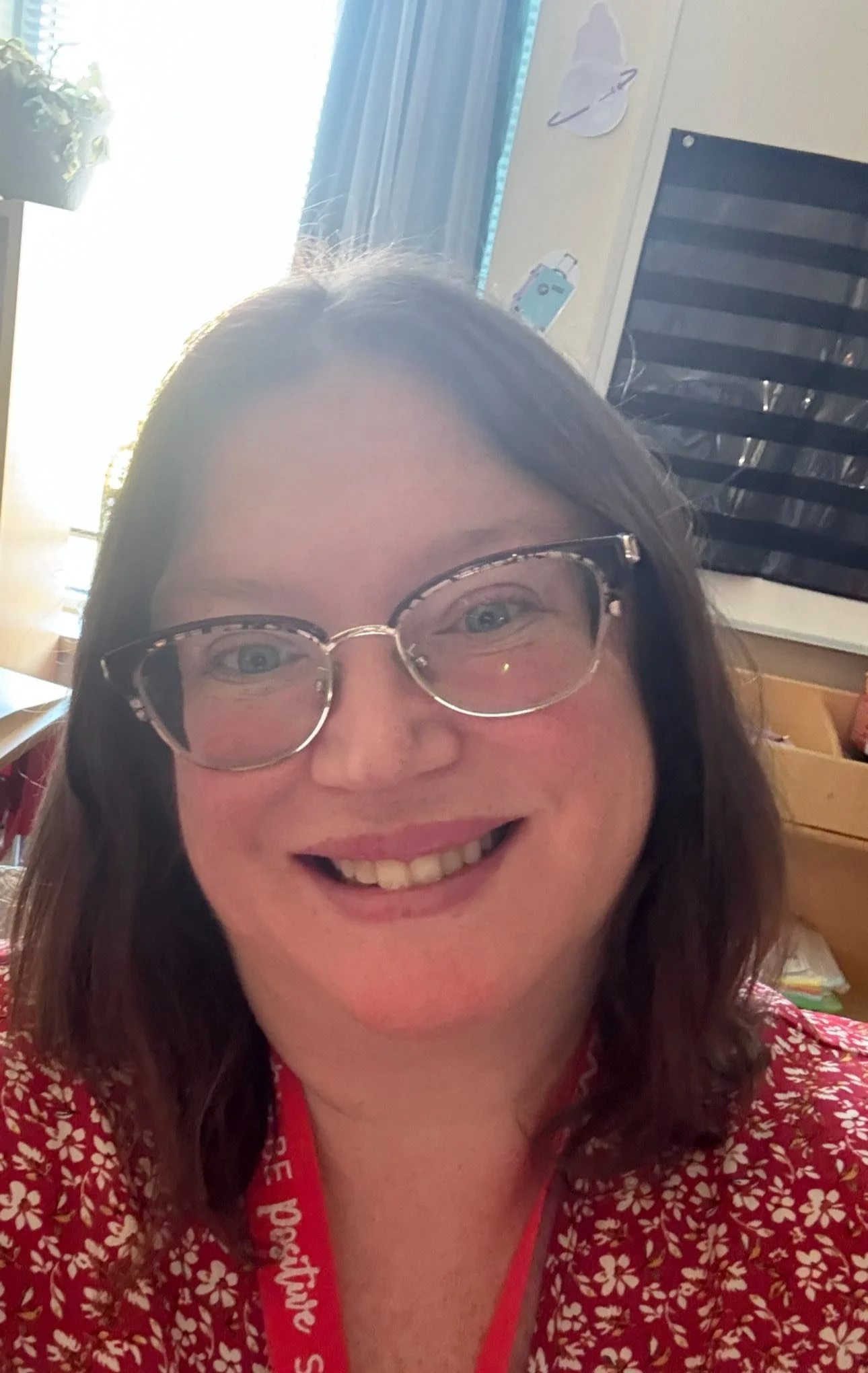 A smiling woman wearing glasses with a patterned top, with a bright window and some decorations on the wall behind her.