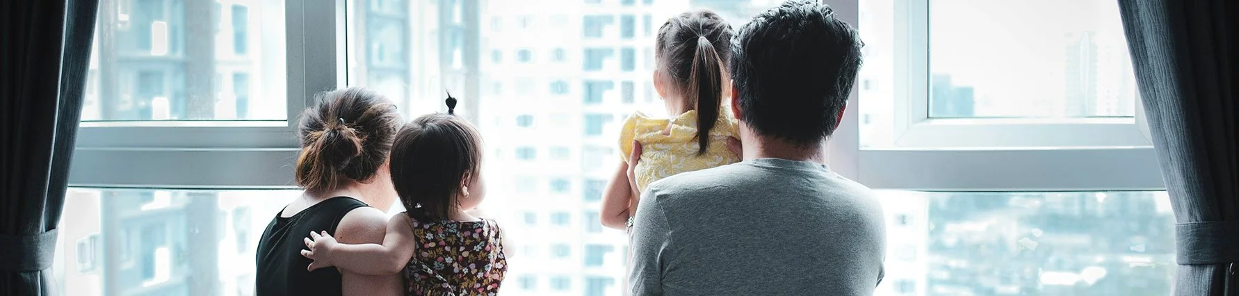 Mom and dad, each holding one kid, looking out a large apartment window together at the city skyline