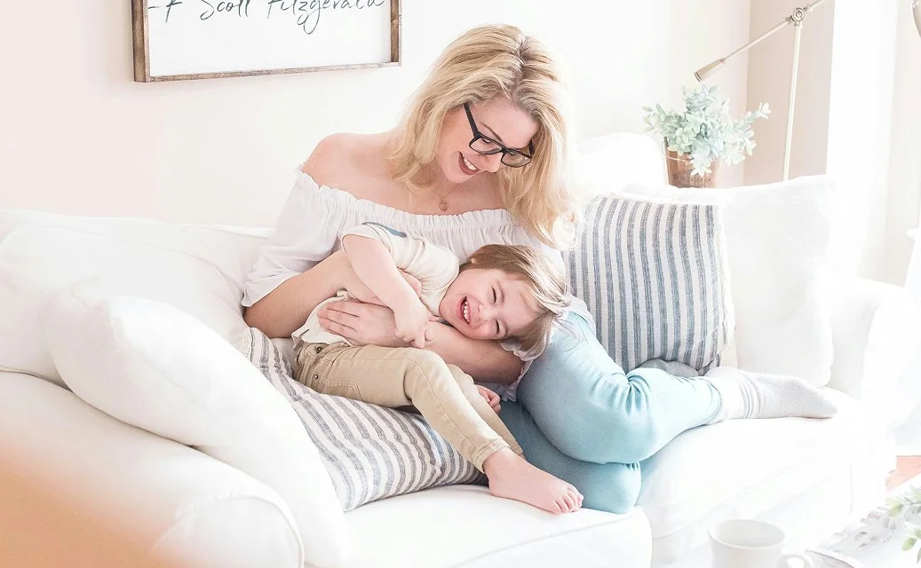 A mother and her child cuddling and laughing together on a couch in a bright, cozy living room