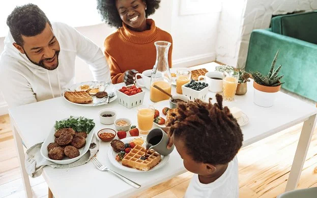Family laughing, gathered together around a breakfast table sharing waffles, fruit, and juice in a bright kitchen dining space