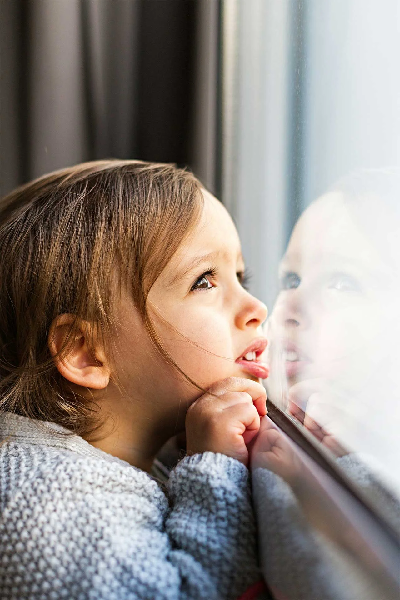 Child looking out a window hopefully, with hands resting on the windowsill and her face reflected in the glass in front of her