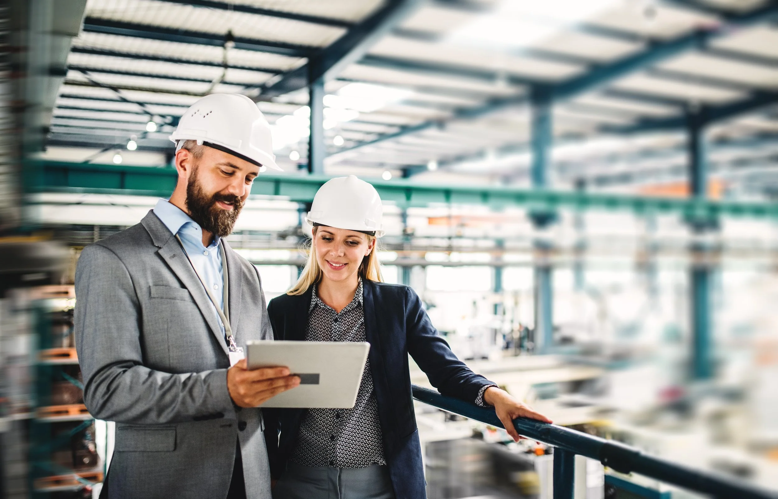 Two professionals, a man and a woman, wearing white safety helmets and business attire, looking at a tablet in an industrial setting with shelves and equipment in the background.