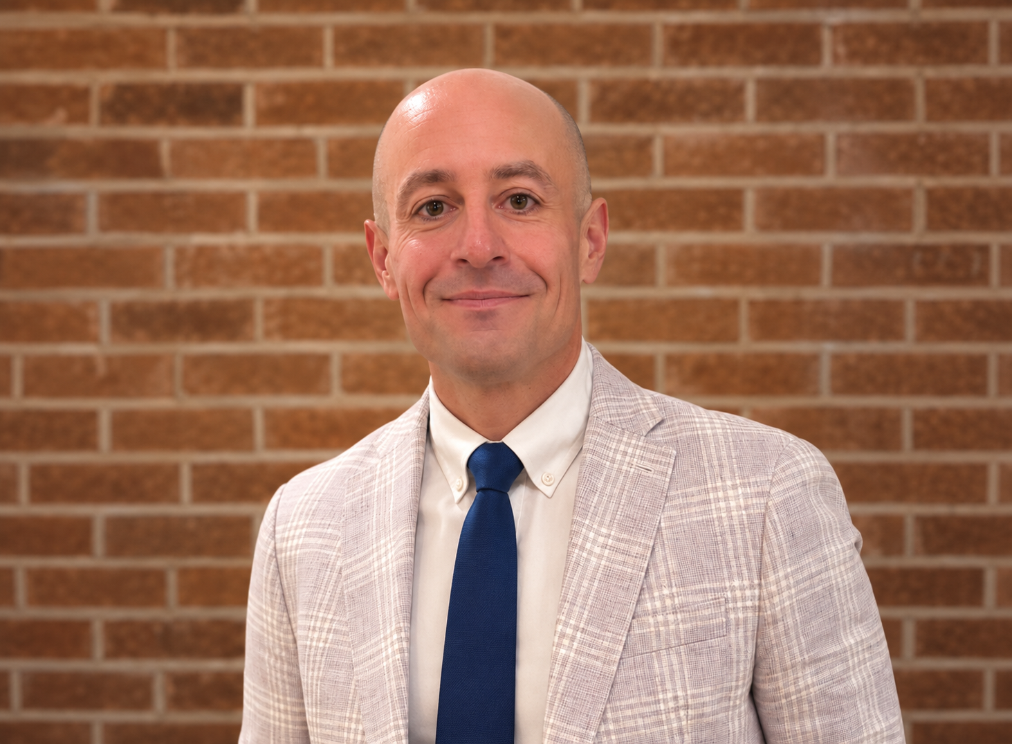 A man with a bald head and light skin, wearing a light-colored plaid suit jacket, a white dress shirt, and a blue tie, standing in front of a brick wall.