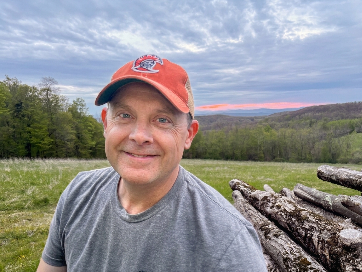 A man smiling outdoors in a grassy field with logs stacked behind him, a forest, and a cloudy sky with hints of pink at sunset in the background.