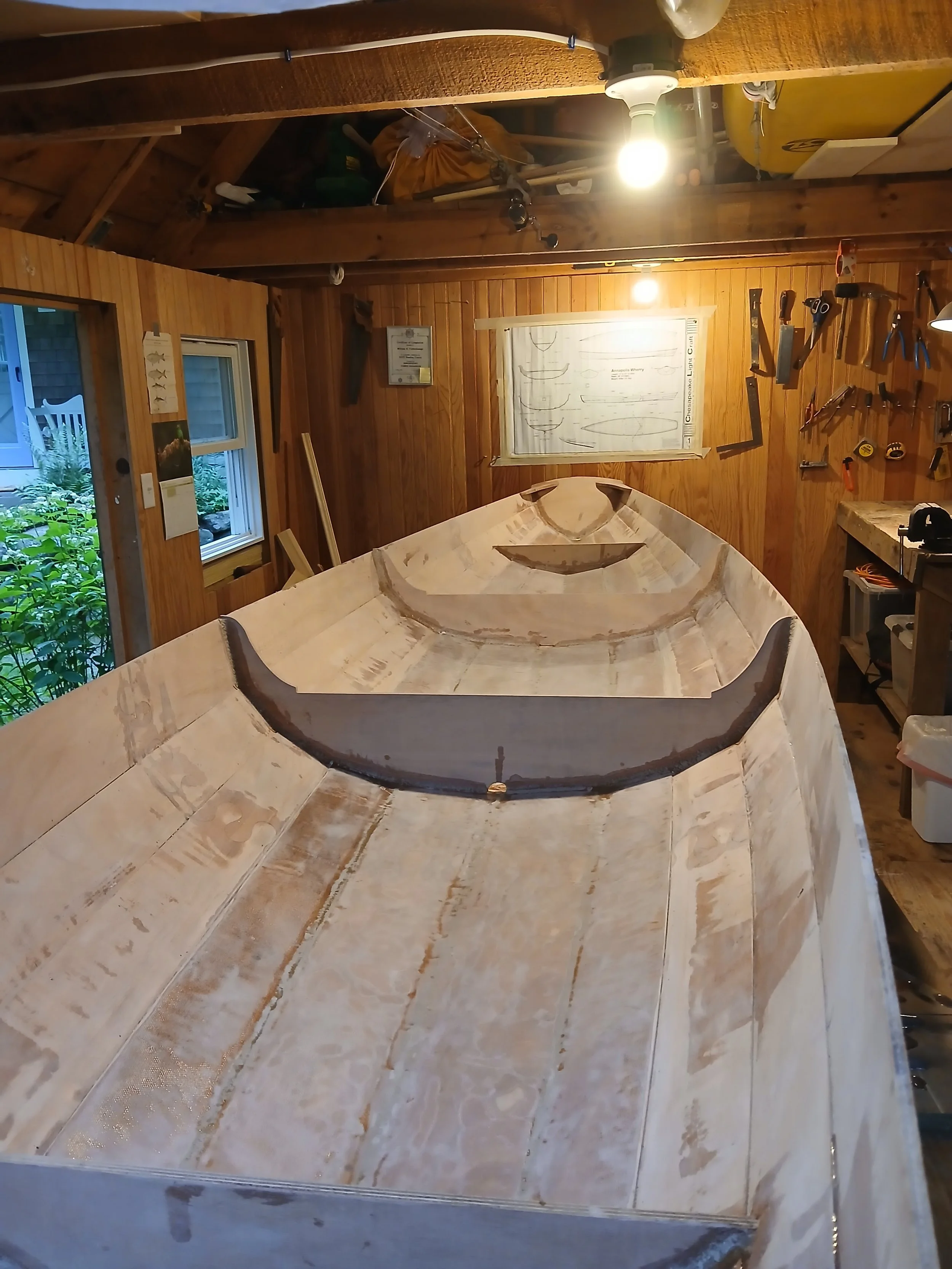 A woodworking workshop with a partially built wooden boat inside. The boat is placed on a workbench, and various tools hang on a wood-paneled wall. A window shows an outdoor scene with greenery.
