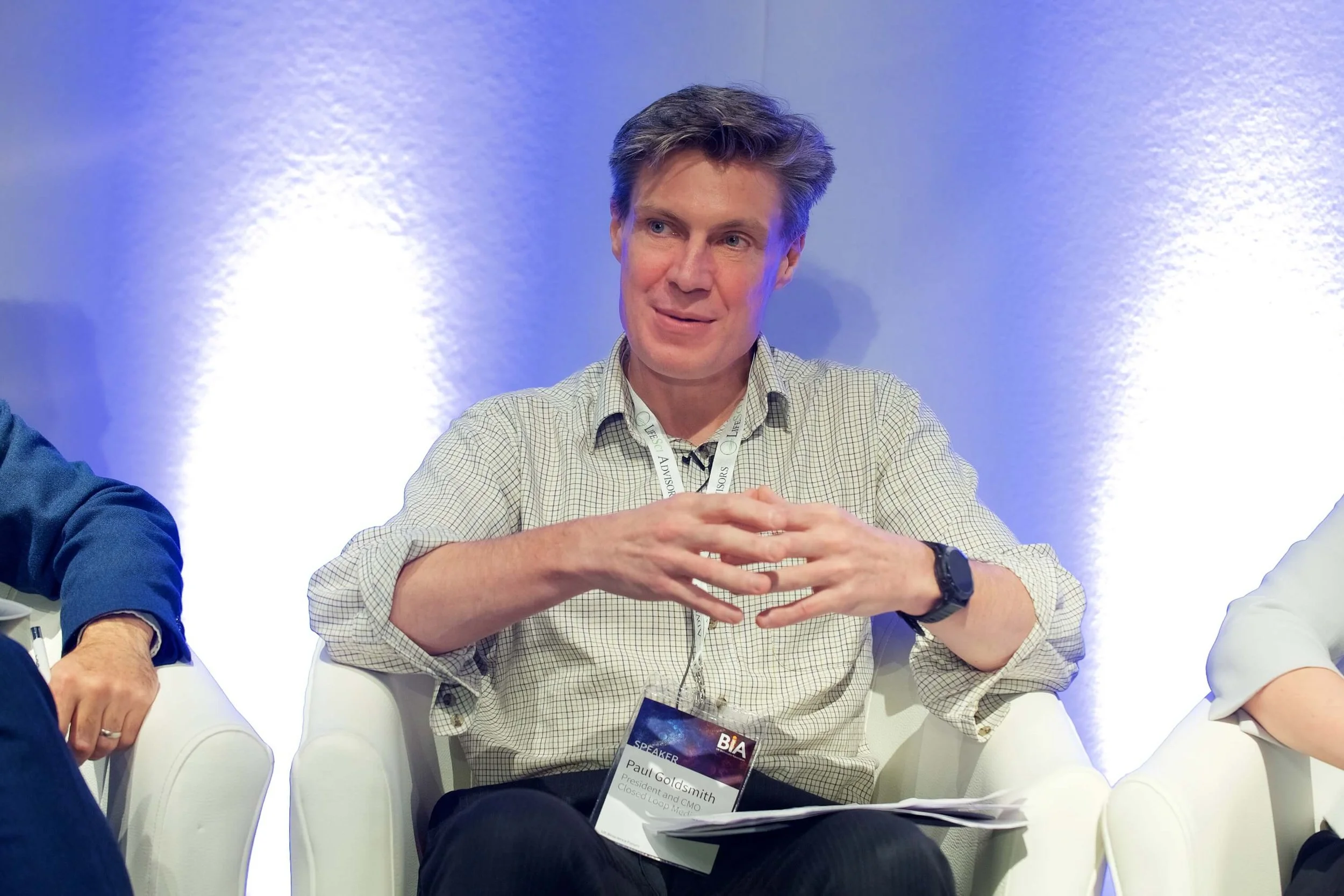A man with brown hair wearing a beige checkered shirt sits at a panel discussion, gesturing with his hands, with a purple-lit background.