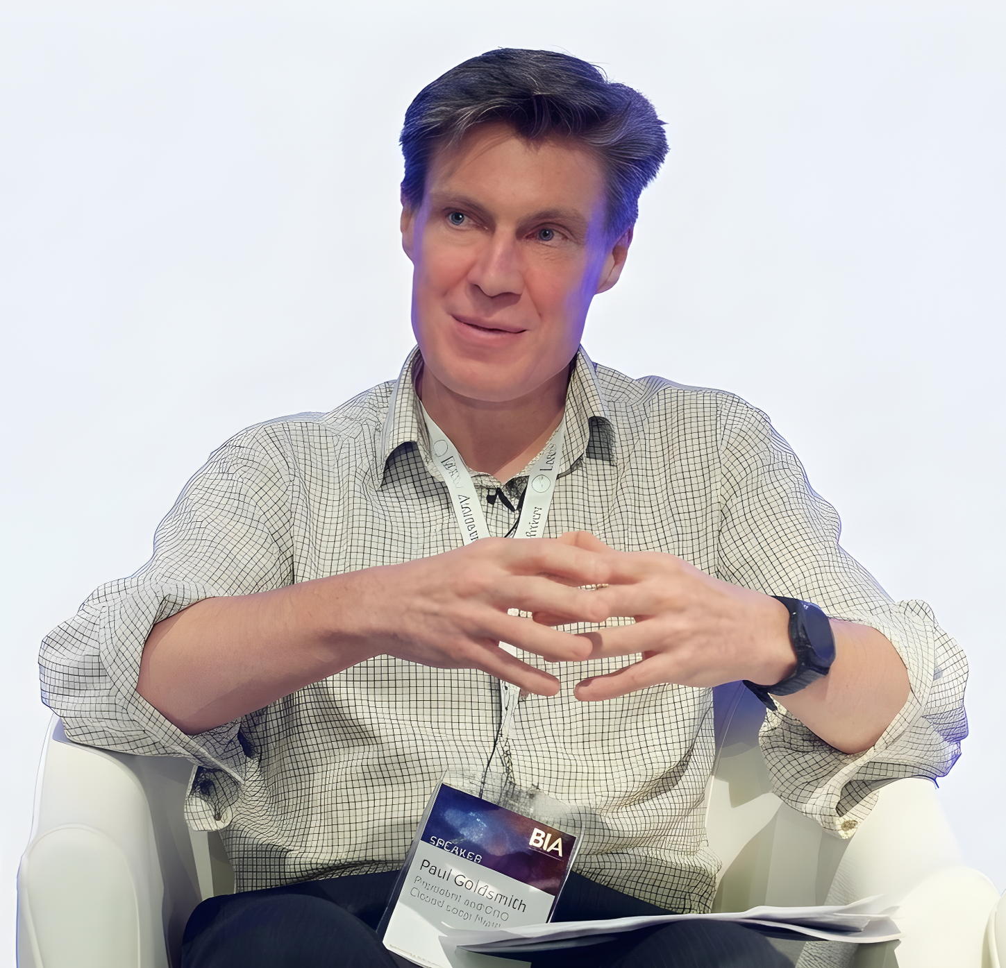 Man with brown hair, wearing a checkered shirt, sitting in a white chair at a conference or event, with a lanyard and name badge, speaking or listening attentively.