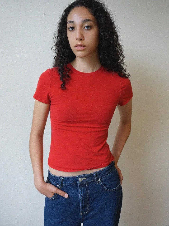 A young woman with long, curly black hair wearing a red t-shirt and blue jeans standing against a plain off-white wall.