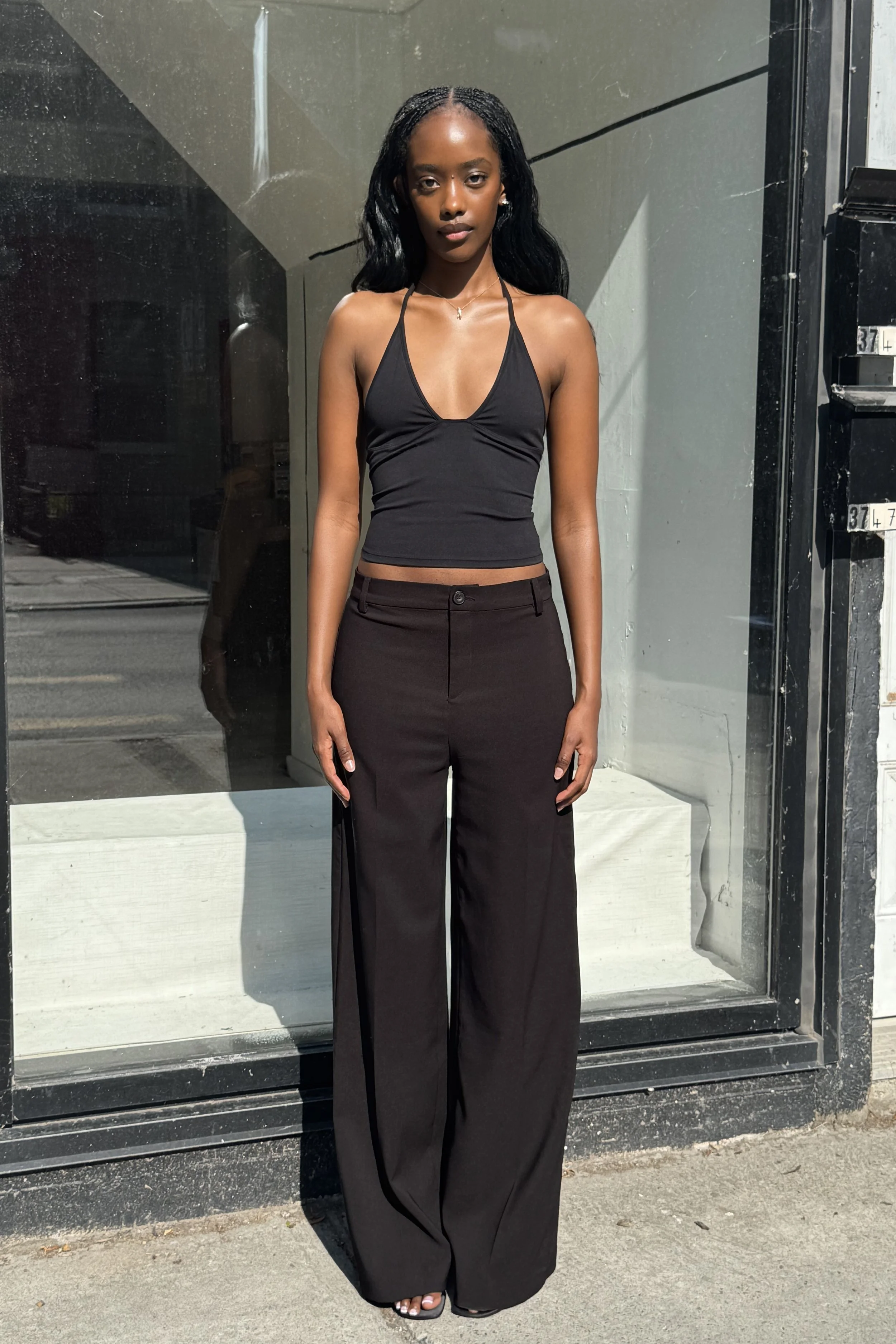A young woman with dark skin and long curly hair stands outside in front of a reflective glass window and black door. She is wearing a black halter top and black wide-leg pants, with open-toed sandals. She looks directly at the camera with a neutral 