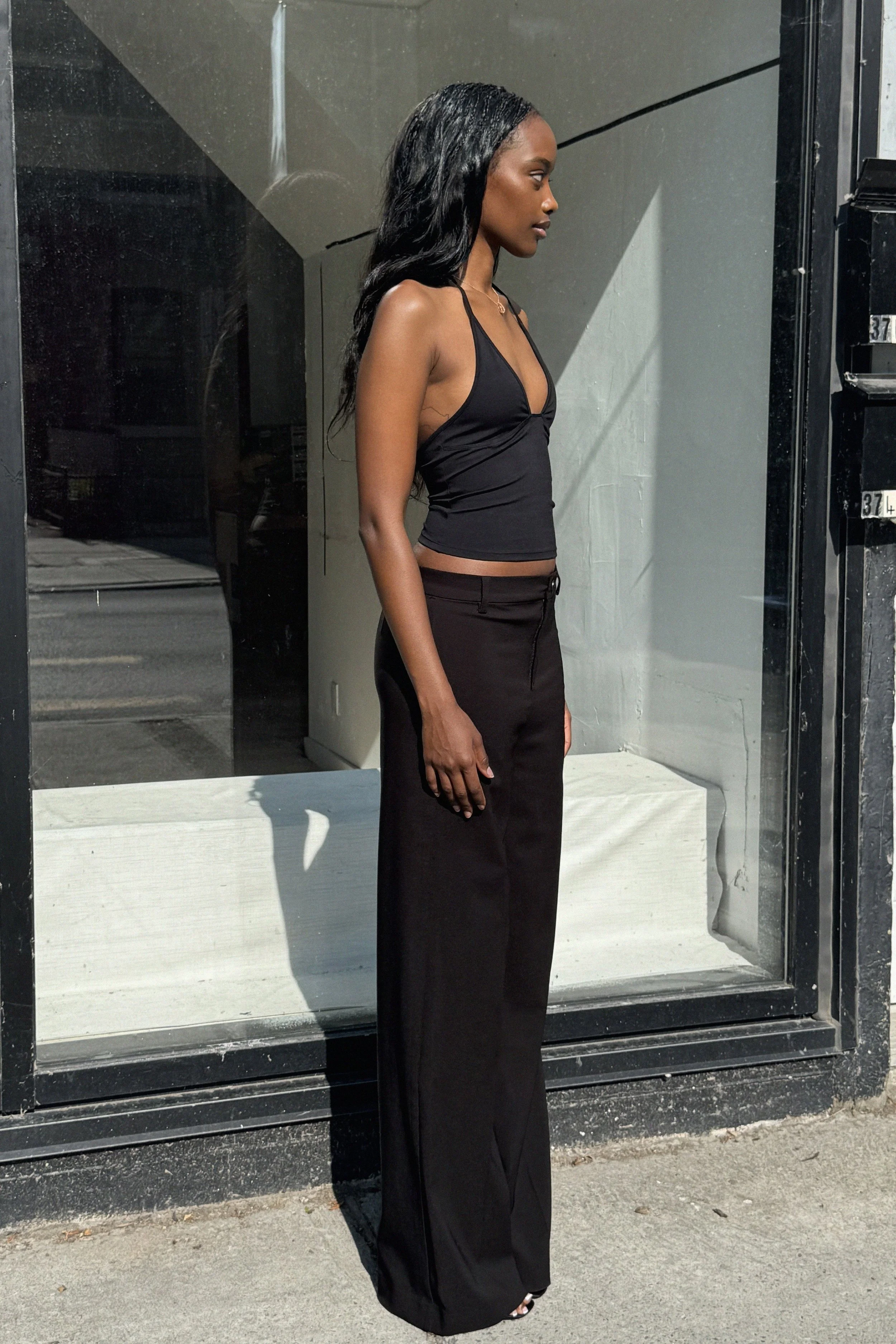 A woman standing outside a store, wearing a black sleeveless top and black wide-leg pants, with long dark wavy hair, in profile view.