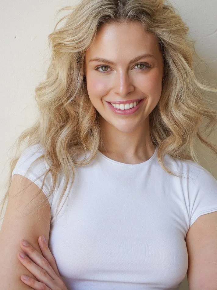Noémi, a young Montréal female model signed with MTL Models MGMT  with blonde, wavy hair smiling at the camera, wearing a white t-shirt, against a plain light-colored wall.
