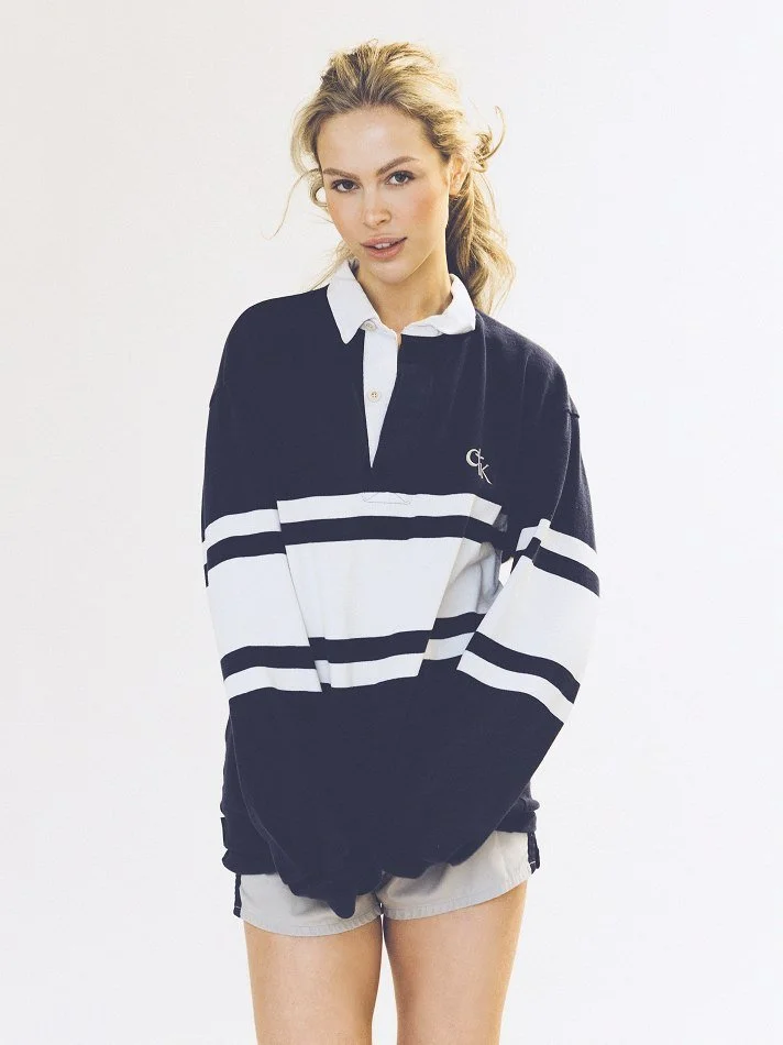 Noémi, a young Montréal female model signed with MTL Models MGMT with blonde hair wearing a black and white striped rugby shirt and beige shorts posing against a plain white background.