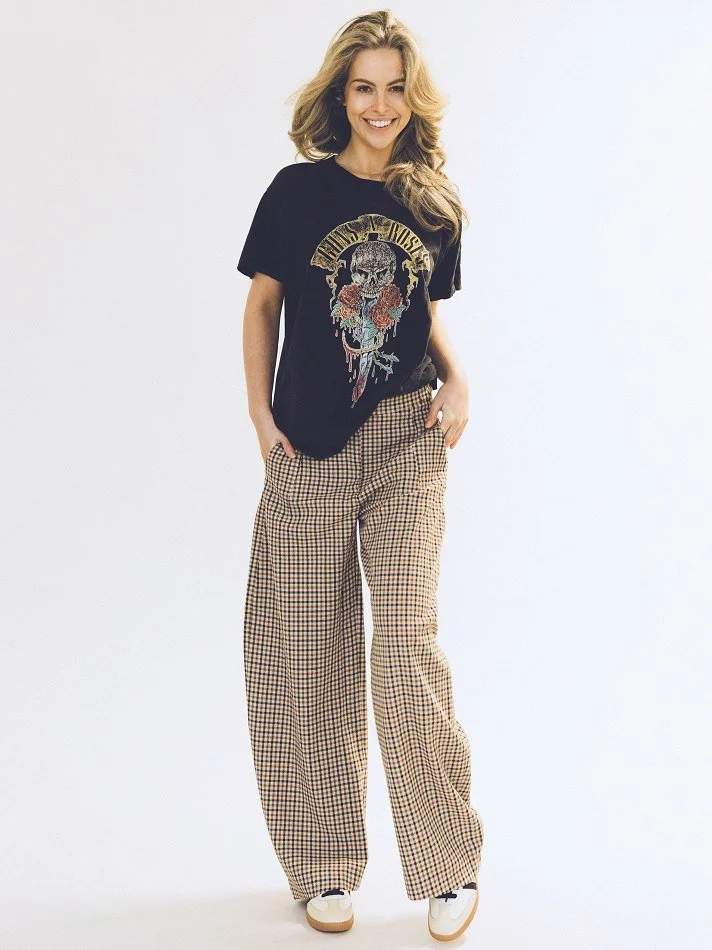 Noémi, a young Montréal female model with long, wavy blonde hair, smiling and standing against a plain white background. She is wearing a black graphic T-shirt with a skull and roses design and checkered wide-leg pants, paired with white sneakers.