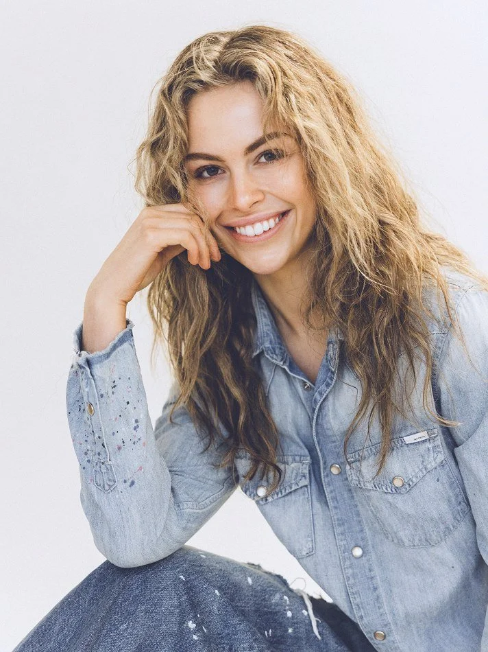 Noémi, a young Montréal female model signed with MTL Models MGMT with wavy blonde hair smiling and resting her chin on her hand, wearing a light denim jacket and modelling sitting against a plain white background.