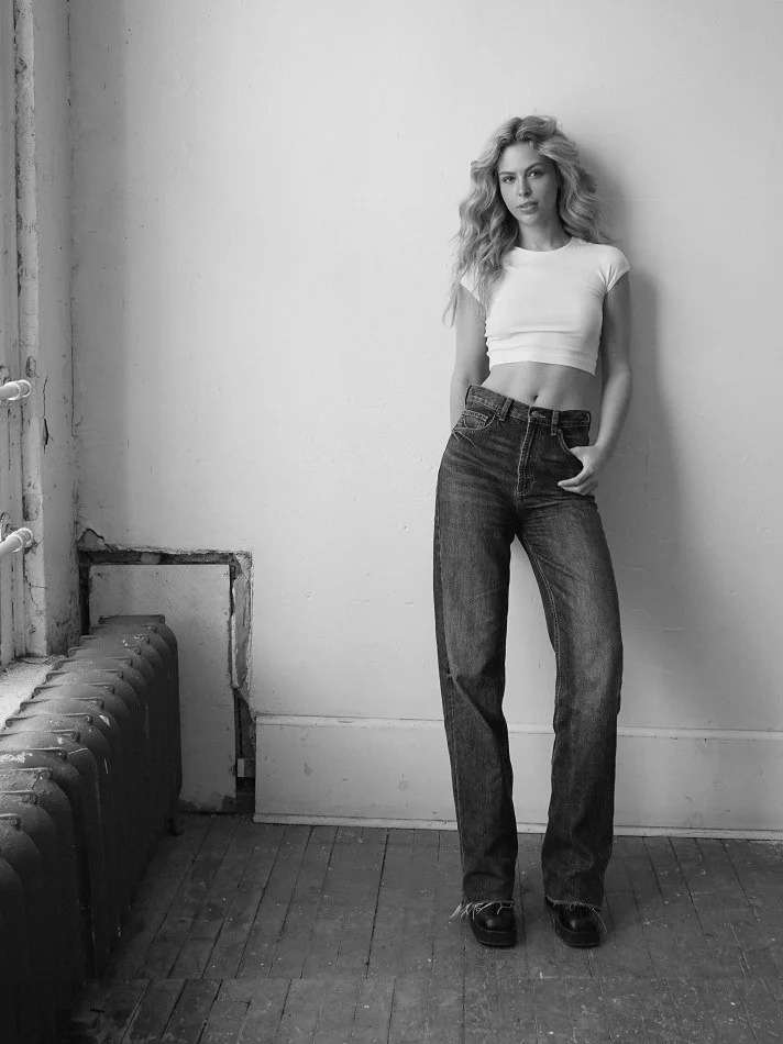 Black and white photo of Noémi, a young Montréal female model signed with MTL Models MGMT with long wavy hair, wearing a cropped white t-shirt, high-waisted loose jeans, and dark shoes, standing against a plain wall.