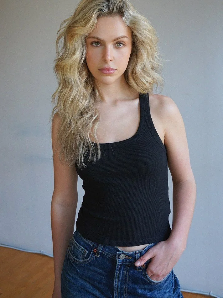 Noémi, a young Montréal female model signed with MTL Models MGMT with long, wavy blonde hair, wearing a black sleeveless top and blue jeans, modelling against a plain light-colored wall.