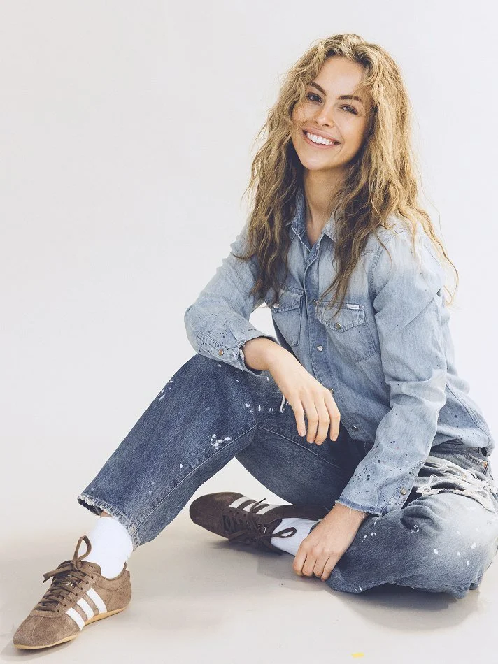 Noémi, a young Montréal female model signed with MTL Models MGMT with curly blonde hair sitting on the floor against a white background, smiling, wearing a denim jacket, jeans with paint splatters, white socks, and brown sneakers.