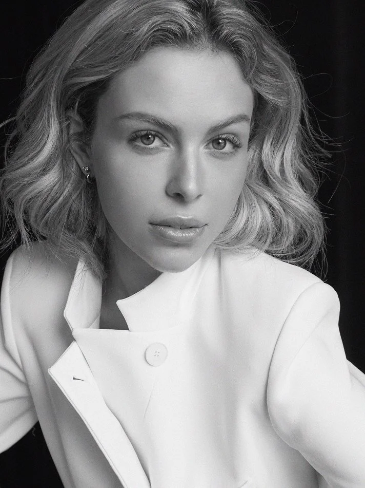 Noémi, a young Montréal female model signed with MTL Models MGMT  with wavy blonde hair looking directly at the camera, wearing a white coat, black background.