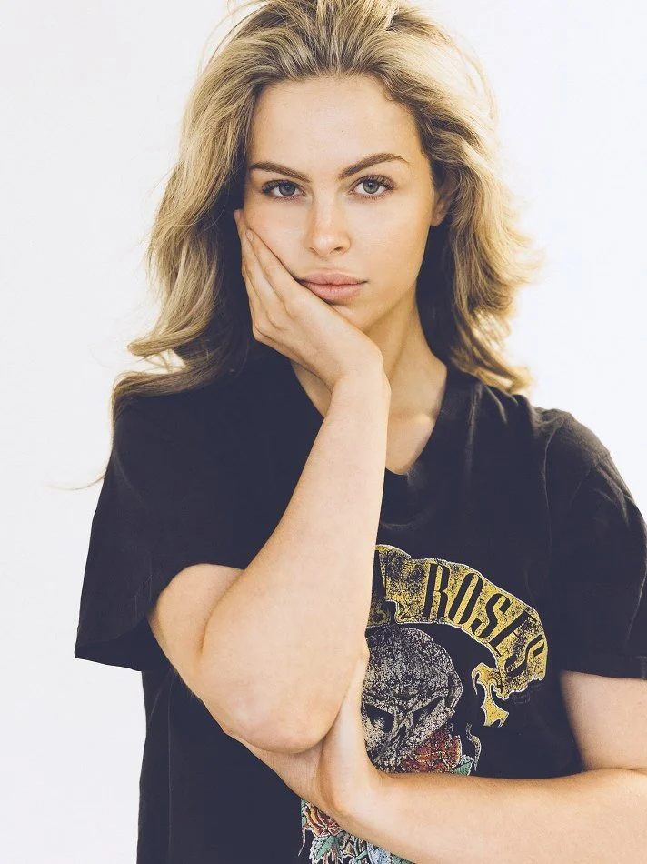 Noémi, a young Montréal female model signed with MTL Models MGMT with wavy blonde hair wearing a black T-shirt with a graphic design, posing with her hand on her face against a plain background.