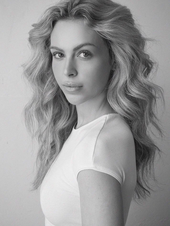 A black-and-white portrait of Noémi, a young Montréal female model signed with MTL Models MGMT with wavy, voluminous hair, wearing a white top, modelling at the camera with a neutral expression.