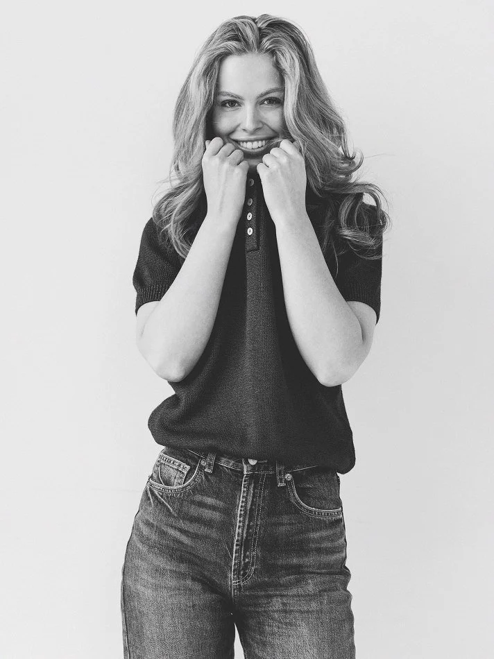 Noémi, a young Montréal female model signed with MTL Models MGMT  with wavy hair, smiling and modelling holding her fists up near her face, dressed in a dark polo shirt and jeans, standing against a plain background.