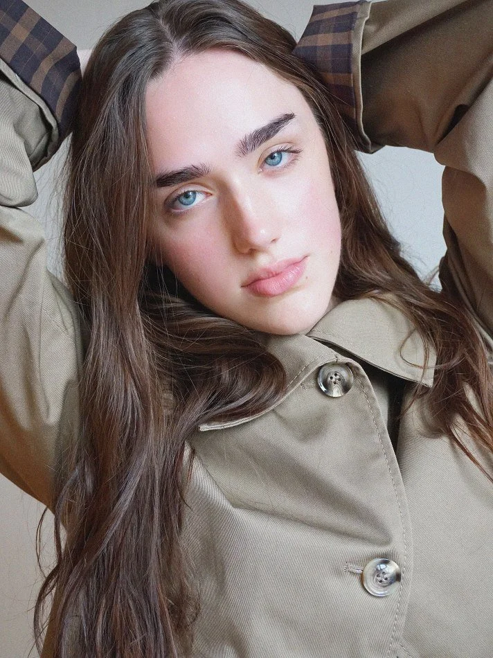 Close-up of Violet, a young Montréal female model signed with MTL Models MGMT with long brown hair and blue eyes, wearing a beige trench coat, modelling looking at the camera with a relaxed expression.