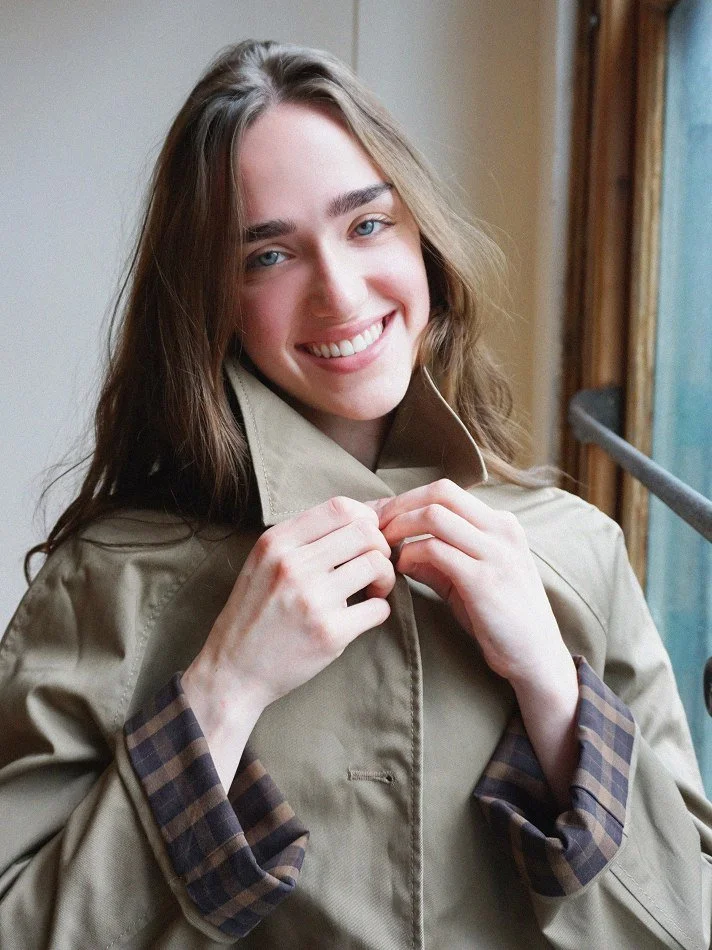 Violet, a young Montréal female model signed with MTL Models MGMT modelling agency, she has long brown hair and blue eyes smiling, adjusting her beige trench coat, modelling near a window with natural light.