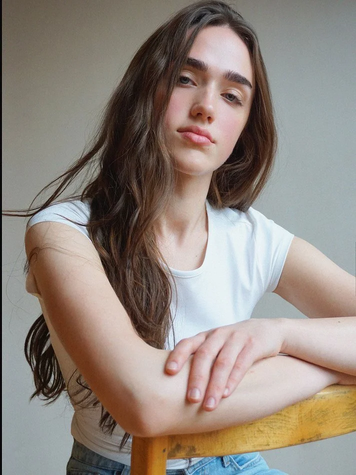 Violet, a young Montréal female model signed with MTL Models MGMT with long brown hair and fair skin sitting at a wooden table, wearing a white t-shirt, looking at the camera with a neutral expression.