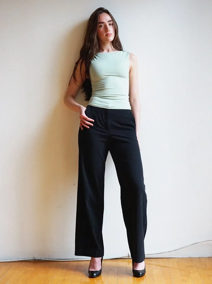 Violet, a young Montréal female model signed with MTL Models MGMT with long brown hair wearing a light green sleeveless top, black wide-leg trousers, and black high heels modelling against a plain off-white wall in a Montreal studio.