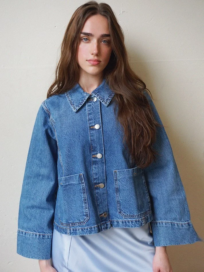 Violet, a young Montréal female model signed with MTL Models MGMT with long brown hair wearing a blue denim jacket and a light-colored skirt modelling against a plain wall in a Montreal studio.