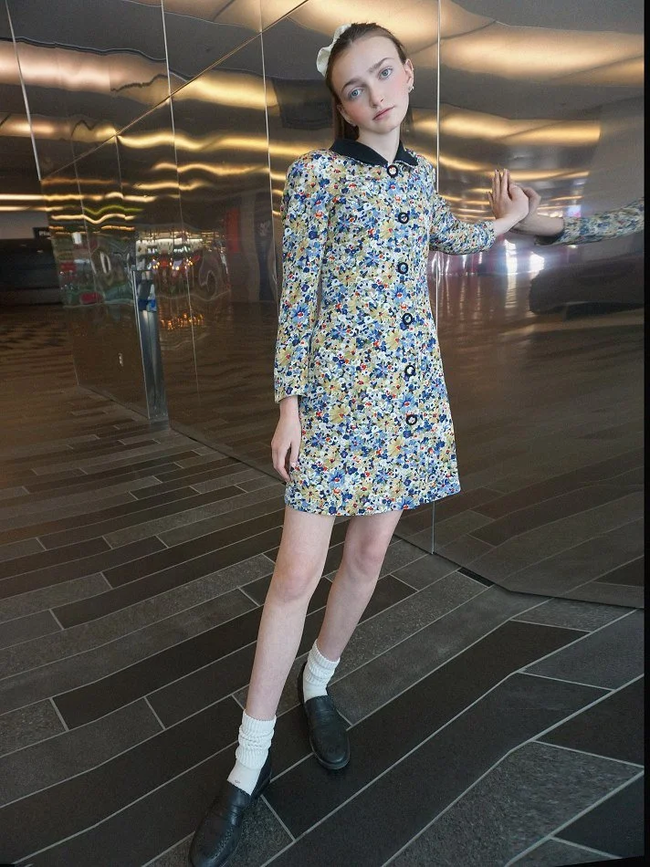 Sadie, a young Montréal female m with long hair and light makeup leaning against a reflective, metallic wall, wearing a floral dress with black collar and buttons, white socks, and black shoes, in an indoor setting with a modern, artistic background.