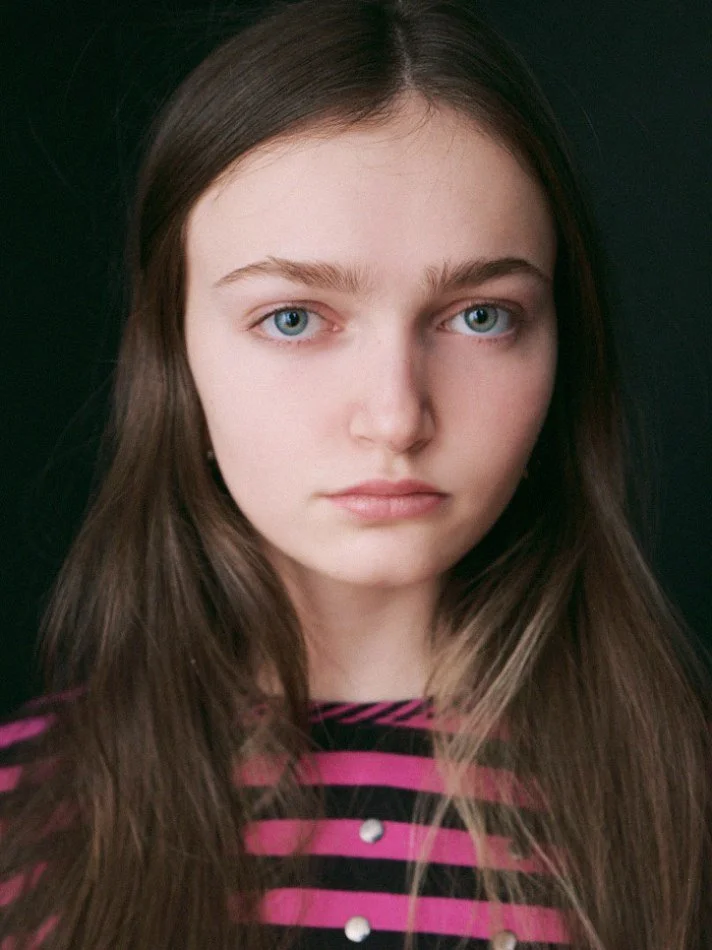 Close-up portrait of Sadie, a young Montréal female model signed with MTL Models MGMT with long brown hair, blue eyes, and a serious expression, wearing a pink and black striped top against a dark background.