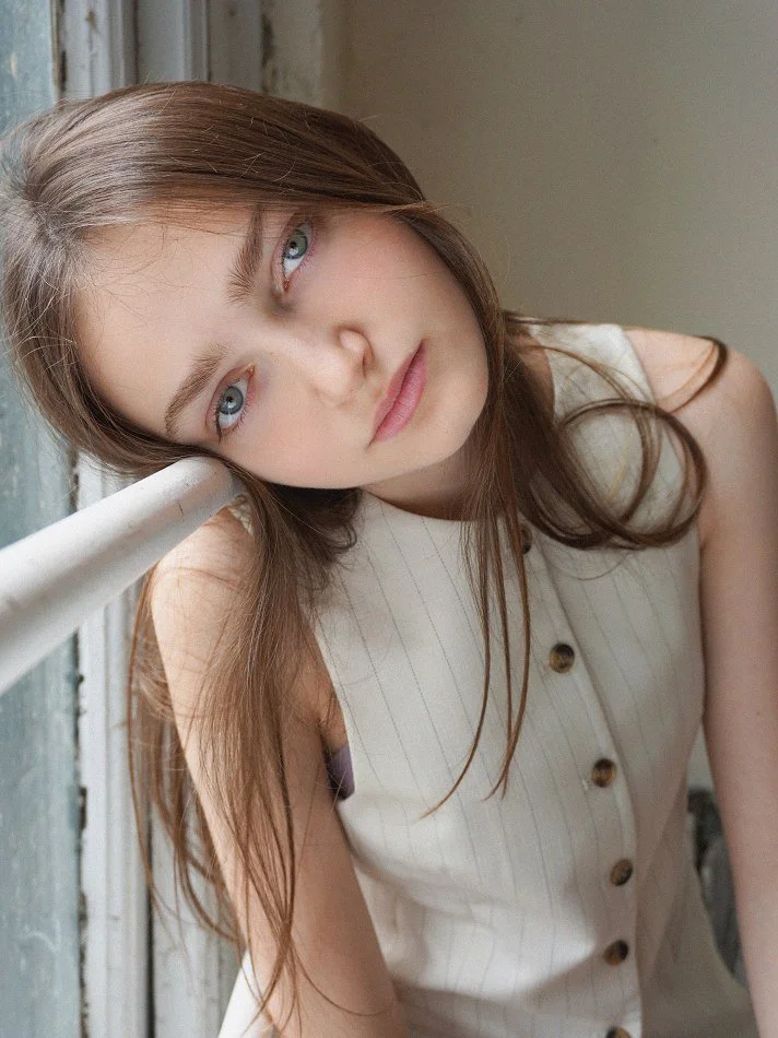 Sadie, a young Montréal female model signed with MTL Models MGMT with long brown hair and blue eyes leans her head on a white railing, posing at the camera. She wears a sleeveless cream-colored top with buttons.