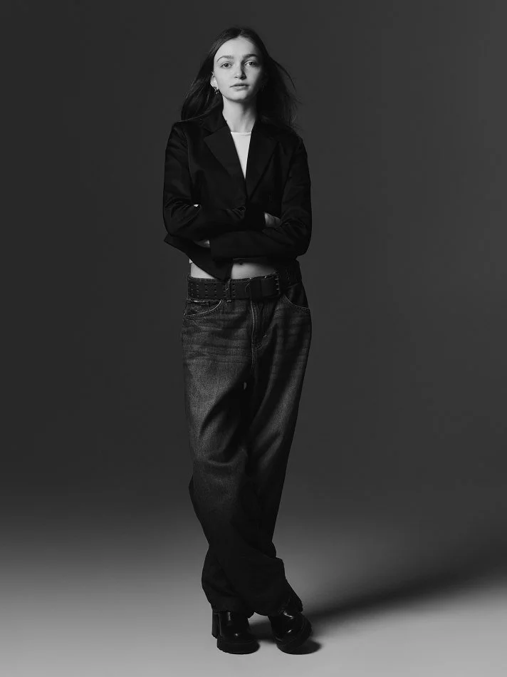 Black and white photograph of Sadie, a young Montréal female model signed with MTL Models standing against a plain gray background. She is wearing a black blazer, high-waisted jeans, and black boots, with her arms crossed and a serious expression.