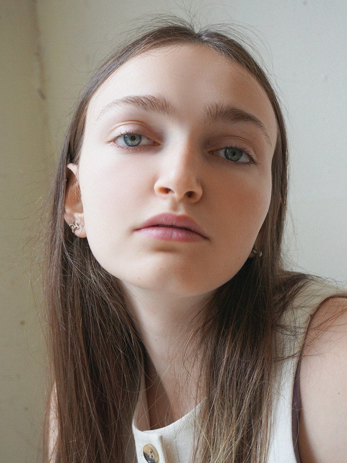 Close-up of Sadie, a young Montréal female model signed with MTL Models MGMT with long brown hair, light skin, and blue eyes, wearing earrings and a sleeveless top, looking into the camera. Shot by photographer Melina Querel.