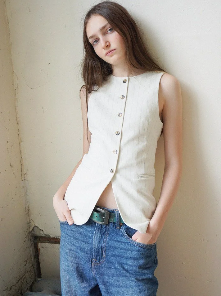 Sadie, a young Montréal female model signed with MTL Models MGMT with long brown hair and blue eyes leaning against a beige wall, wearing a sleeveless white vest with vertical pinstripes and button details, paired with blue jeans.