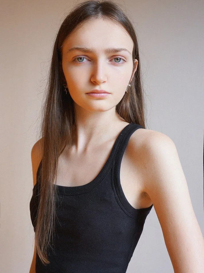 Sadie, a young Montréal female model signed with MTL Models MGMT with long brown hair and blue eyes wearing a black tank top, looking directly at the camera against a plain background. Shot by Montreal photographer Melina Querel.